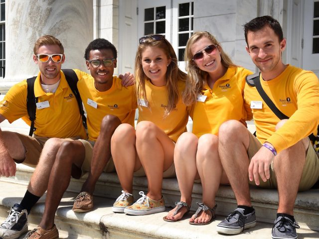 four orientation staff students wearing yellow shirts sit on steps with arms around each other