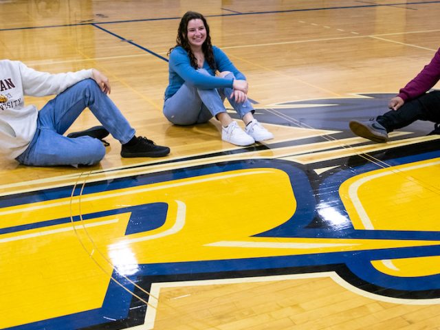 Rowan students sit on a gymnasium floor at a community college, showing the school's logo.