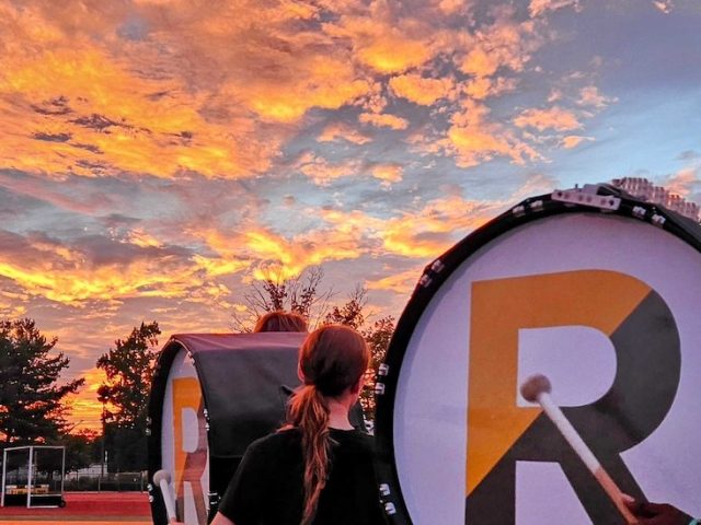 A close up of the Rowan University marching band drum major practicing on the athletic field with a dramatic sunset in front of him.