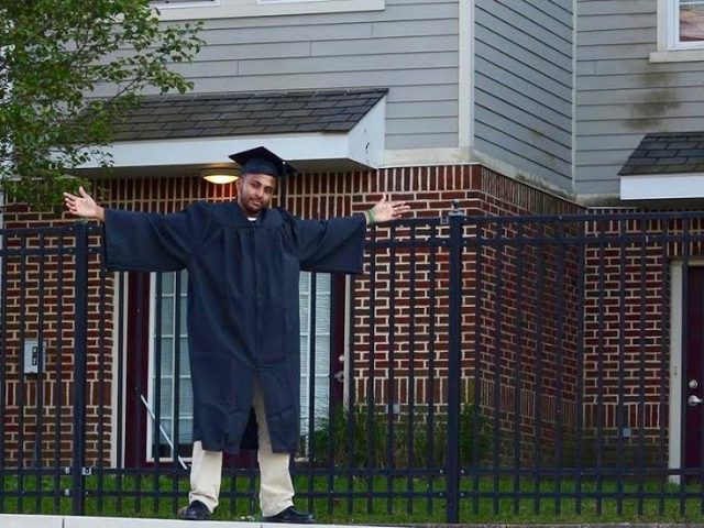 Sha stands in his cap and gown with arms outstretched on top of a Rowan sign