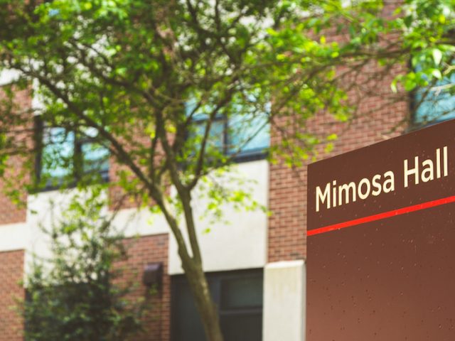 brown sign for Mimosa Hall, in front of building with blooming green trees