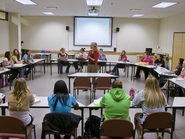 A classroom with tables set up in the shape of a circle with a professor and lots of students