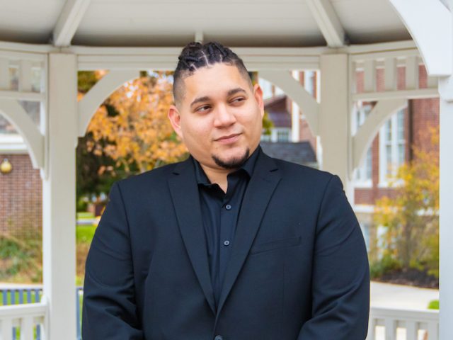 Angel wears a dark blue suite inside the white gazebo on campus