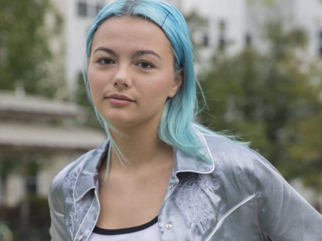 Rowan University student with blue hair wearing a silver silk jacket standing outside Robo