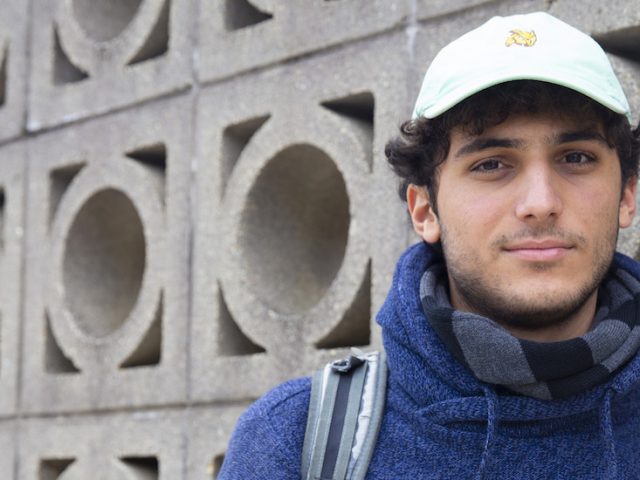 Jacks stands in front of a a wall with a circular concrete pattern, wearing a blue sweatshirt, backpack and Rowan hat