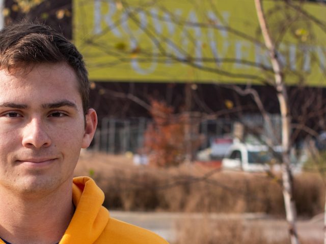 Young, white male student with yellow Rowan University standing in foreground with brick building blurred out in background