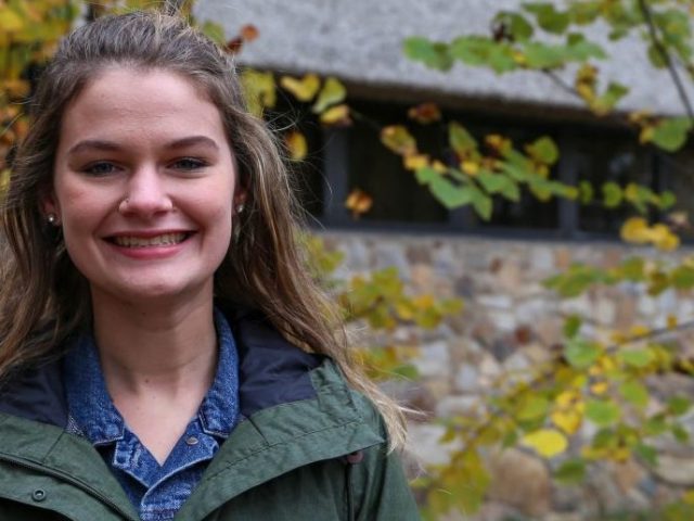 Sarah stands in front of autumn trees outside of Robinson Hall, smiling and wearing a green jacket