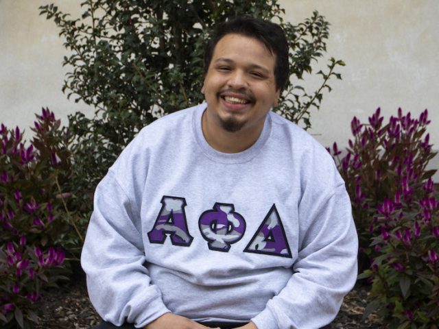 Victor sits on the brick area outside the student center while wearing a Greek Alpha Phi Delta sweatshirt