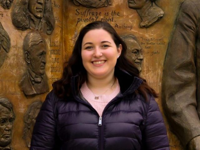 Rebecca Shnier stands outside of James Hall at Rowan University, in front of a bronze artwork that says Knowledge is Power