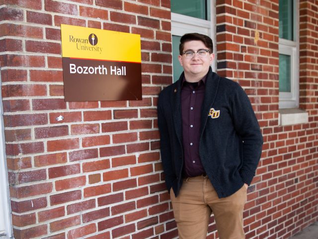 Brandon outside Bozarth Hall in Rowan sweater, in front of brick wall