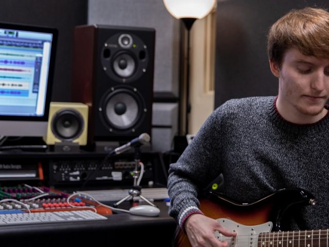 Young male student playing electric guitar inside a music production student while sitting down in a chair