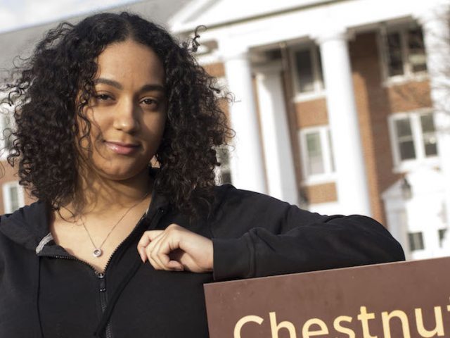 Vanessa from Rowan University stands in front of the sign for Chestnut Hall dorm, with her left elbow resting on the brown sign