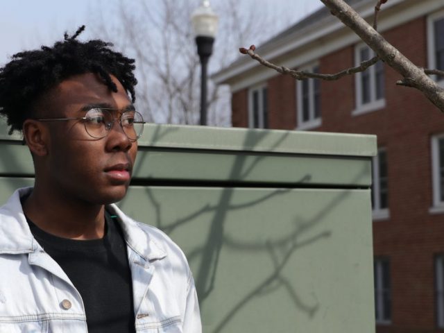 Jelani wears a ripped white denim jacket over a black t-shirt, standing in front of a pale green metal wall near dorms at Rowan University