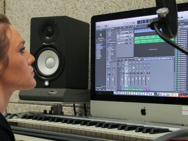 Claire sits at a table with a Mac computer and microphone to work on a song in Wilson Hall's private recording studio.