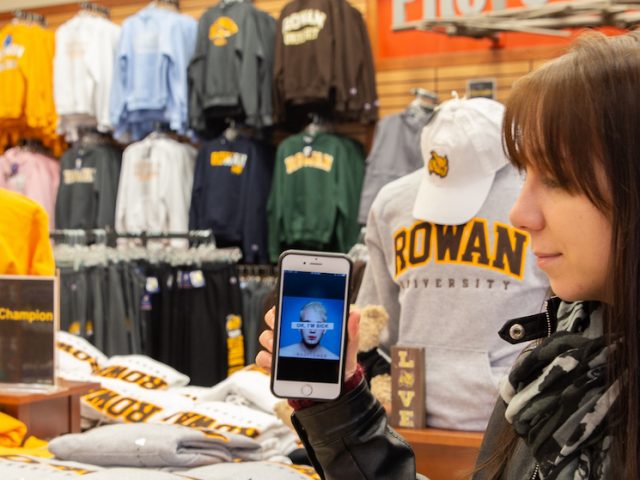 Jen Green stands in the Rowan University bookstore, looking toward her phone with the Badflower album cover OK I'm Sick