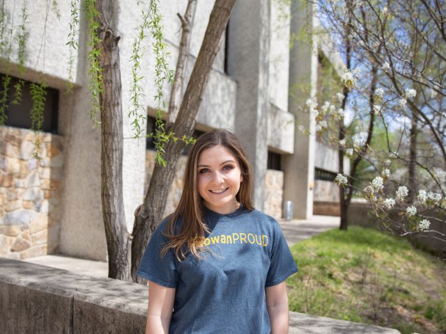 A Rowan University student wearing a #RowanProud shirt stands outside James Hall