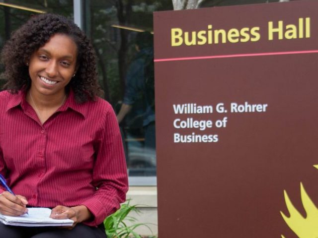 Jo Carter sits next to a Business Hall sign at Rowan University, wearing a pink button down blouse and holding a notebook