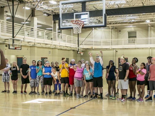 student wearing red shorts and blue shirt makes a basketball shot as his friends watch.