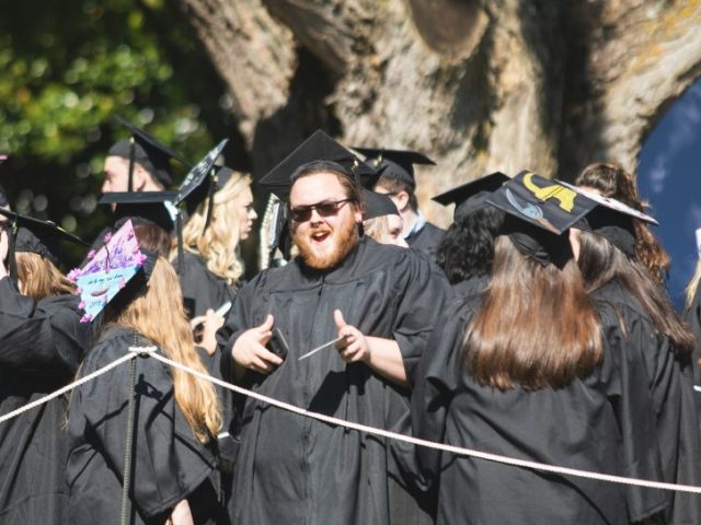 Johnathan Puglise smiles at the camera while being surrounded by other recent graduates in their black cap and gowns.