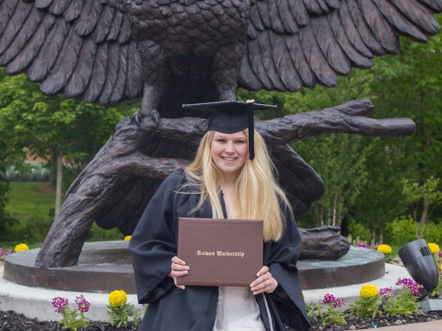 Cheyenne wears her graduation gown, posing with Rowan University diploma holder in front of owl statue