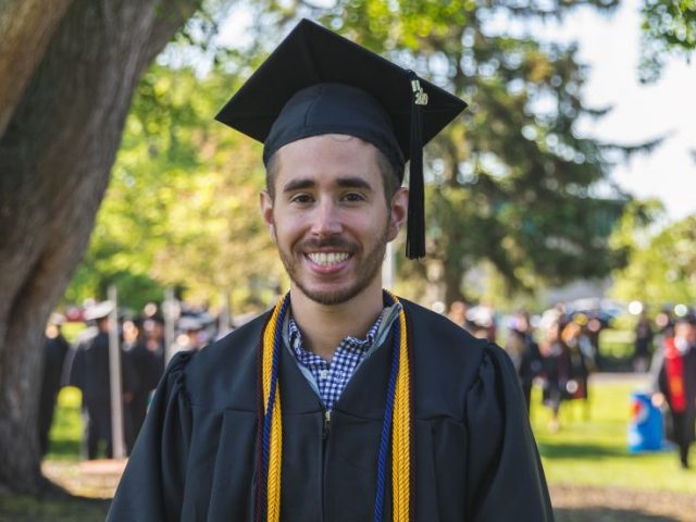 Alex, wearing his graduation cap and gown, standing on the Bunce Green.