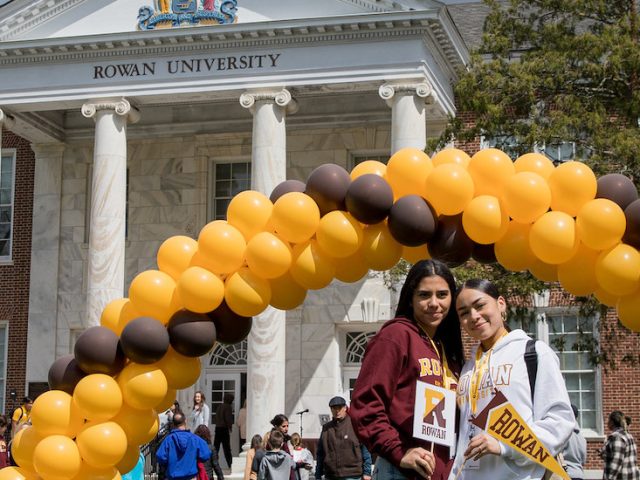 Two young women stand under a brown and gold school colors balloon arch at Rowan University at Accepted Students Day.