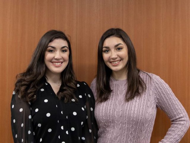 Sisters Sophia and Madison Agostini pose in front of a wood panel wall.