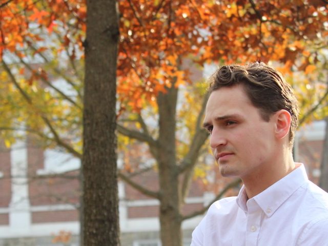 Adam looks into the distance, while wearing a crisp white button down shirt and fall colored trees behind him.