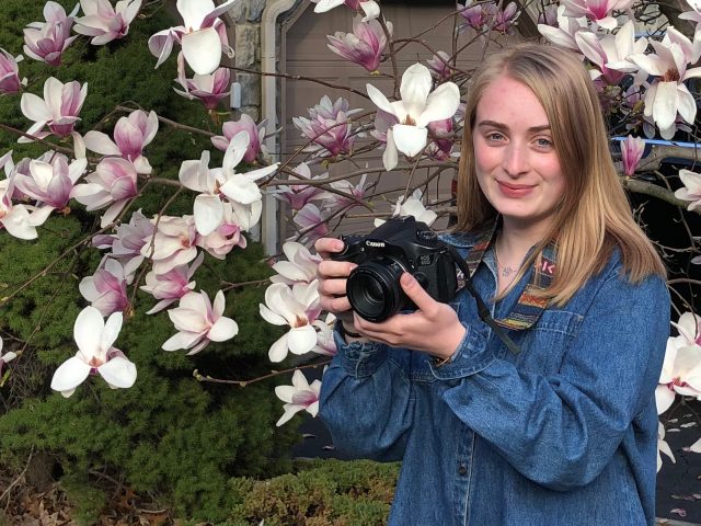 Nicole stands in front of a magnolia tree with a camera.