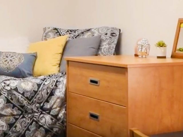 dorm room with gray comforter on bed and wooden desk and drawers.