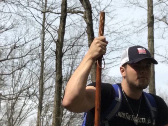 Eddie crouches in the woods, holding a walking stick.