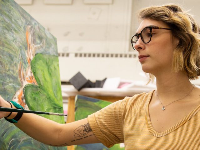 Jessica wears a yellow shirt while holding a paintbrush to her canvas, a nature painting of koi fish.