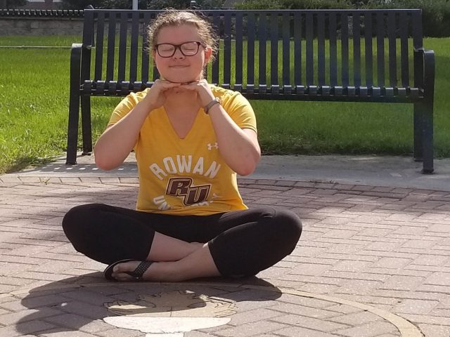 Amanda wears a Rowan shirt and sits on the ground smiling in front of a Rowan logo on the ground.