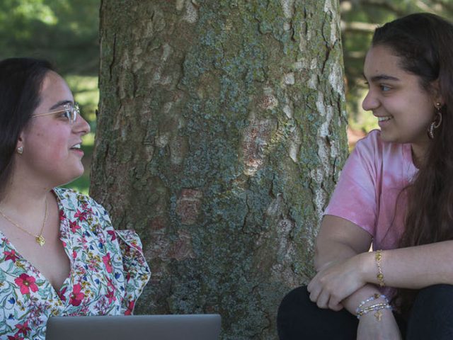 Marlo Rossi sits by a tree with her computer speaking to another student