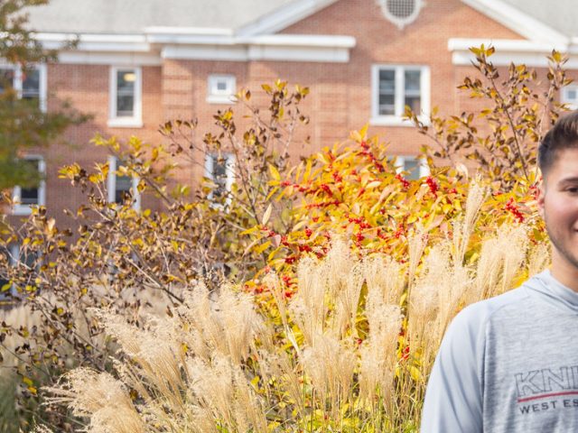 Christopher standing in front of Chestnut Hall. There is green and white shrubbery around him. Trees with green and orange leaves.