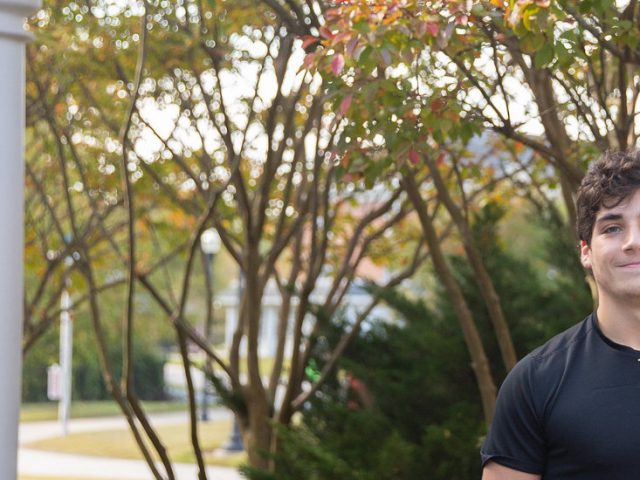 Noah Garcia smiles in front of trees with orange and green. A white building is close by on the left hand side.