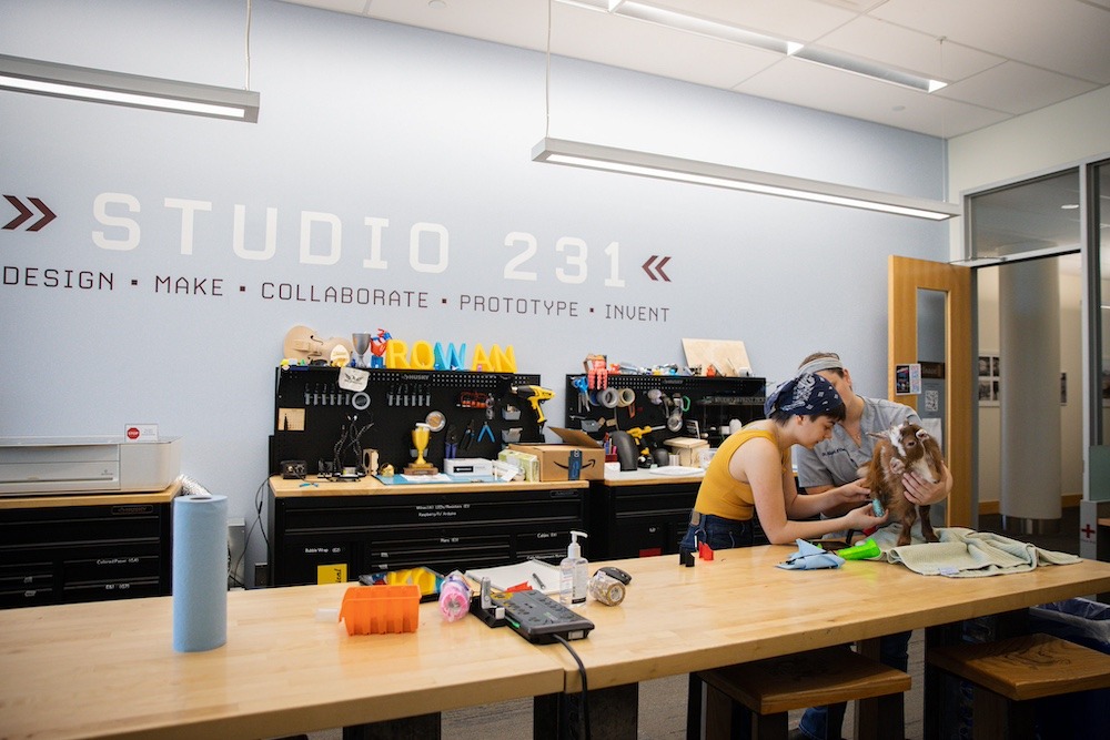 Two Rowan University students hold and hunch over a goat they are fitting for prosthetic feet, while in the makerspace Studio 231.