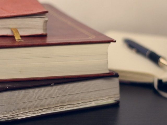 A stack of books and an open notepad with a pen.