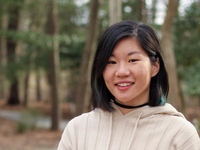 Jean stands outside in front of a wooded area on campus.