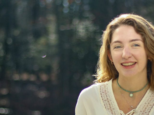 Sarah poses and smiles in front of a wooded area on campus.