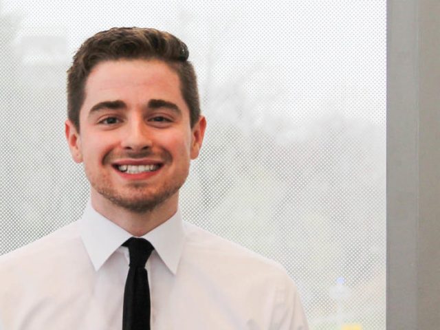 Brett smiles, stands in front of a shaded window on campus.