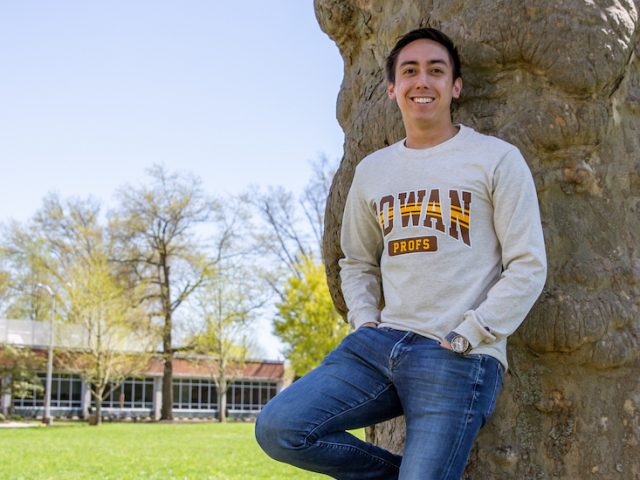 JT leans against a tree on Bunce green, wearing a Rowan sweater with hands in his pockets.