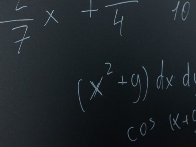 Stock image of math equations being written on a blackboard with chalk in hand.