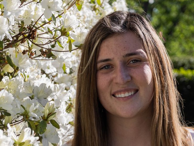 Hannah smiles while standing next to a white flowering plant on campus.