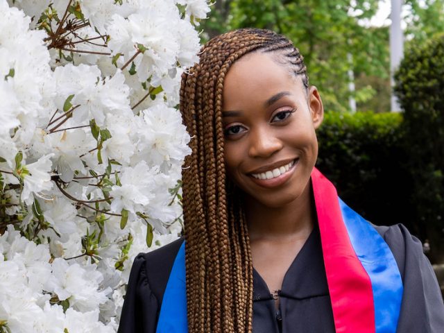 Esther wears her graduation gown and stands in front of campus greenery.