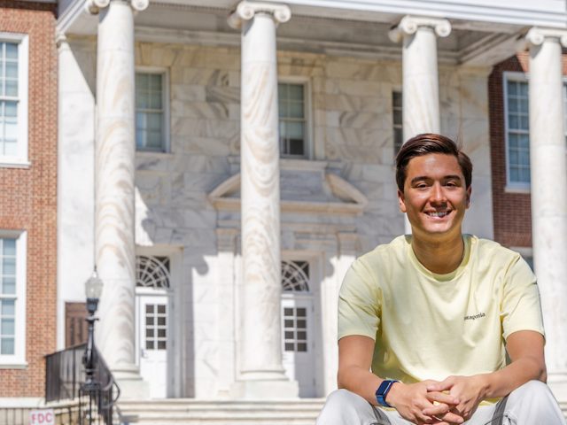 Anthony sits on the grass in front of Bunce Hall wearing a yellow shirt.