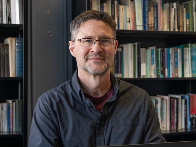 Joe Coulombe sits in front of a stack of bookcases.