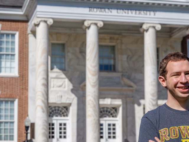 Kevin smiles wearing a gray Rowan t-shirt with Bunce Hall in the distance.