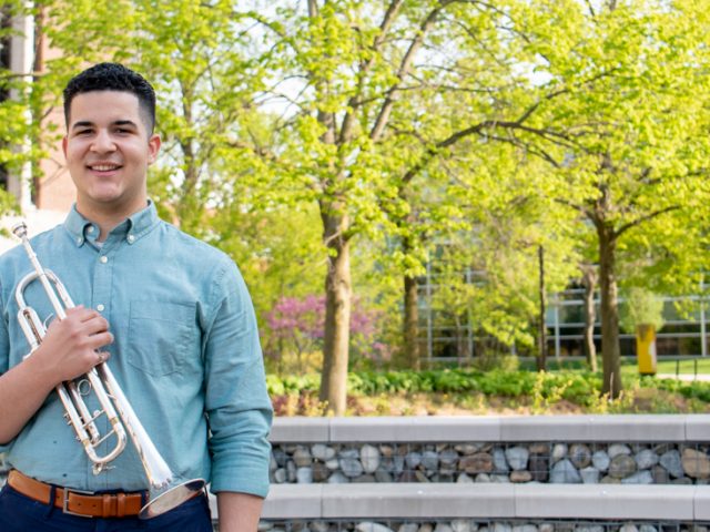 Luis stands with his trumpet outside on campus.