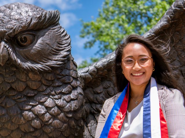 Stephanie sits at the Giant Prof statue on campus while wearing a custom stole showing the Filipino and French flags.